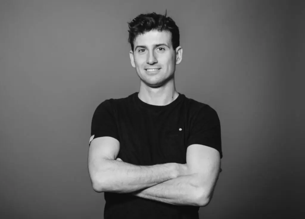 Schwarz-weiß Porträt eines lächelnden Mannes mit kurzen dunklen Haaren, der mit verschränkten Armen vor einem neutralen grauen Hintergrund steht.